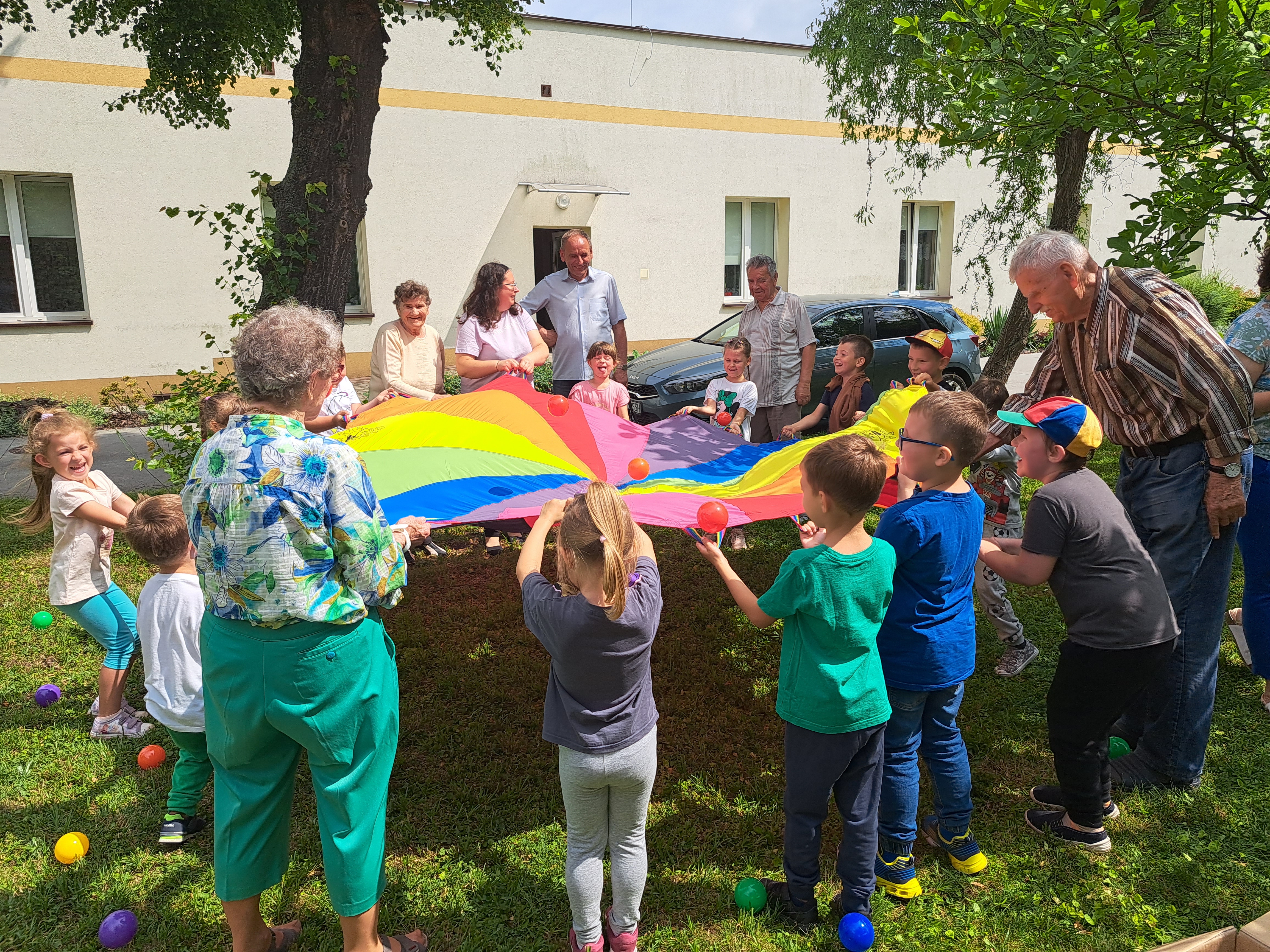 Seniorzy bawią się z dziećmi na podwórku, trzymają kolorową płachtę i próbują wrzucić piłeczkę do środka materiału